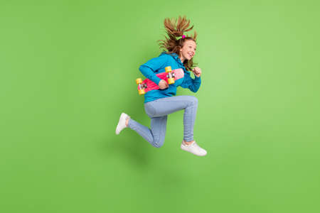 Full body photo of brunette tail haired little girl jump run empty space wear street style outfit longboard isolated on green color backgroundの写真素材