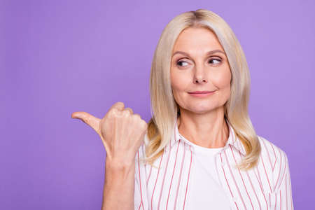 Photo of nice charming mature woman dressed pink shirt pointing thumb up looking empty space isolated purple color backgroundの写真素材