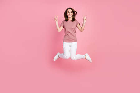 Full length body size view of attractive cheerful girl jumping showing v-sign isolated over pink...