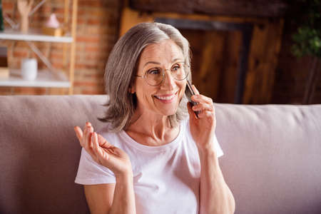 Photo of cute charming lady pensioner wear white t-shirt sitting sofa smiling speaking modern gadget...