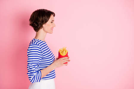 Profile side photo of young beautiful smiling cheerful girl look copyspace hold french fries isolated on pink color backgroundの写真素材
