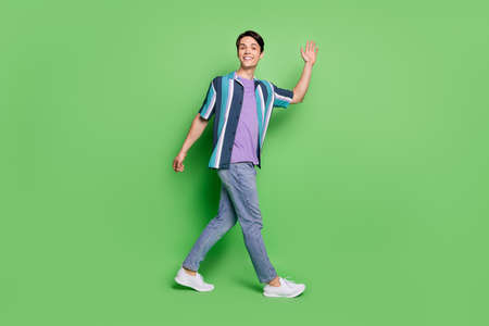 Full body photo of cheerful young happy man wave hello good mood smile walk isolated on green color...