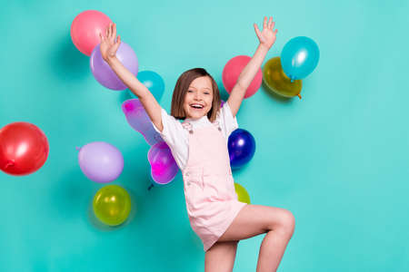 Photo of funny adorable school girl wear pink overall wings smiling playing balloons rising arms isolated teal color backgroundの写真素材