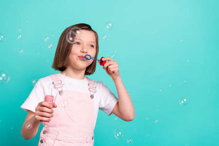 Photo of cute charming schoolgirl dressed pink overall blowing water bubbles smiling isolated turquoise color backgroundの写真素材