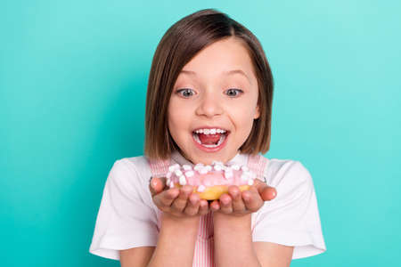 Photo of pretty hungry school girl wear pink overall smiling eating tasty doughnut isolated teal...