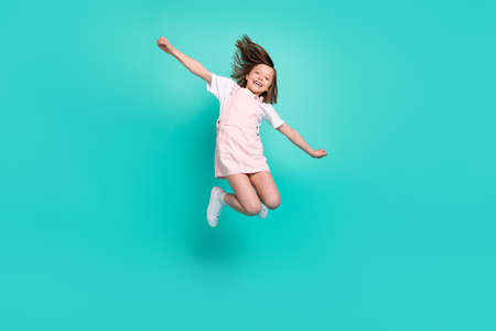 Photo of charming sweet school girl wear pink overall smiling jumping high arms sides isolated teal color backgroundの写真素材