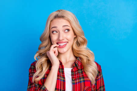 Photo of sweet tricky young woman dressed checkered shirt smiling biting finger looking empty space isolated blue color backgroundの写真素材