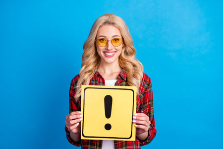 Photo of pretty shiny young lady wear plaid shirt holding exclamation point board isolated blue color backgroundの写真素材