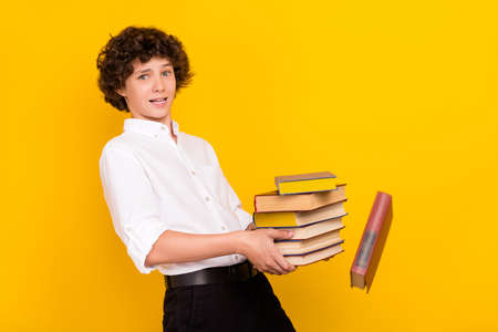Photo of tired upset schoolboy dressed white shirt walking holding many book isolated yellow color background.の写真素材