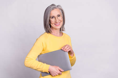 Photo of happy good mood cheerful mature woman in glasses hold laptop wear yellow sweater isolated on grey color backgroundの写真素材
