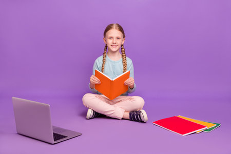 Photo of cheerful positive learner girl sit floor read book toothy smile wear blue shirt isolated purple color backgroundの写真素材