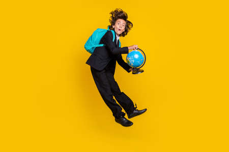 Photo of shocked surprised little boy jump hold globe wear rucksack black uniform isolated yellow...