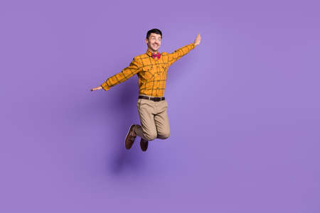 Full length photo of funky carefree young man wear plaid shirt bow tie jumping arms sides isolated violet color backgroundの写真素材
