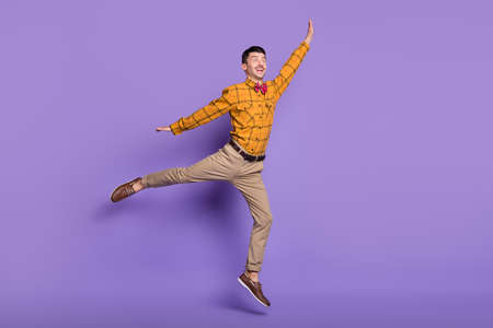 Full length photo of cute sweet young guy dressed checkered shirt jumping arms sides isolated purple...