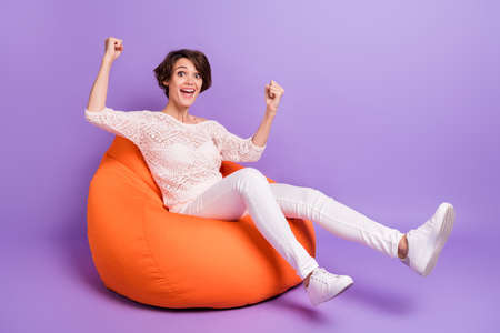 Photo of pretty lucky young woman wear knitted shirt sitting beanbag rising fists isolated violet color backgroundの写真素材