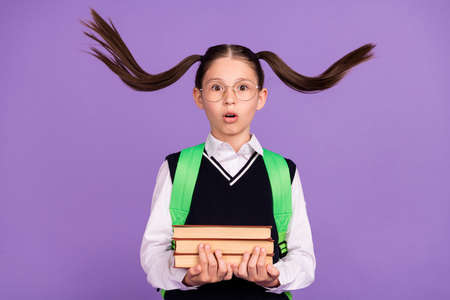 Photo of speechless schoolgirl open mouth hold pile book wear bag glasses uniform isolated purple...