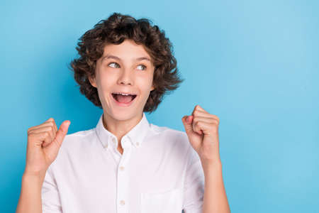 Photo of funny lucky school boy wear white shirt glasses rising fists looking empty space smiling isolated blue color backgroundの写真素材