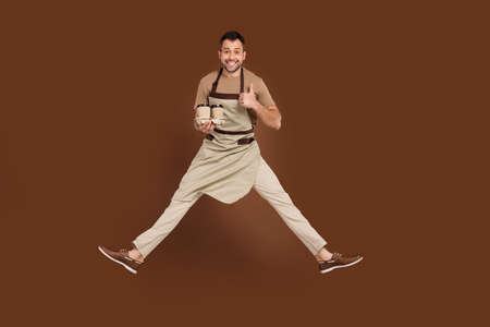 Photo of confident sweet young guy dressed beige uniform jumping showing thumb up preparing latte...