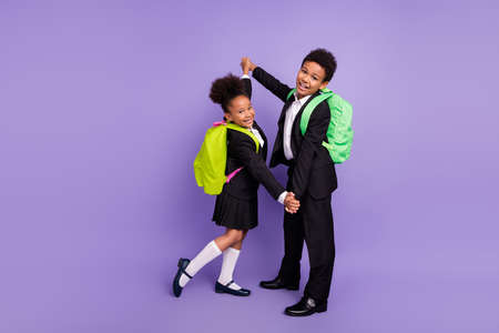 Photo of two african people schoolchildren dance hold hands wear rucksack uniform isolated violet...