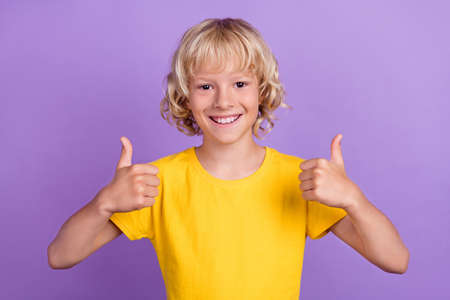 Photo of cheerful positive blond boy raise thumb up approve tips wear yellow t-shirt isolated purple...