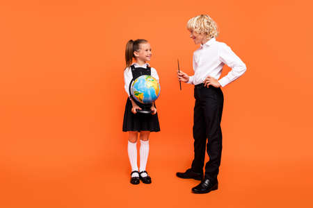 Photo of cheerful schoolchildren hold globe pointer prepare homework wear uniform isolated orange color backgroundの写真素材