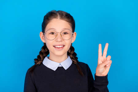 Photo of smiling good mood beautiful clever schoolgirl showing v-sign wear eyewear isolated on blue...