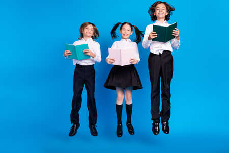Photo of carefree schoolkids jump read homework literature wear uniform shoes isolated blue color backgroundの写真素材