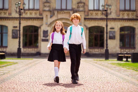 Photo of two schoolkids walk street hold hands enjoy trip wear backpack white shirt uniform park outdoorsの写真素材