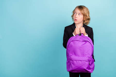 Photo of impressed small blond boy look empty space wear bag uniform isolated on blue color backgroundの写真素材