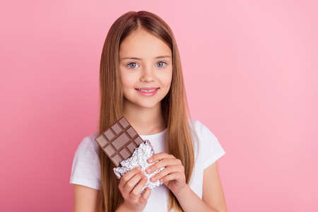 Photo of lovely blond little girl eat chocolate wear white t-shirt isolated on pink color backgroundの写真素材