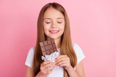Photo of nice blond small girl eat chocolate closed eyes wear white t-shirt isolated on pink color backgroundの写真素材