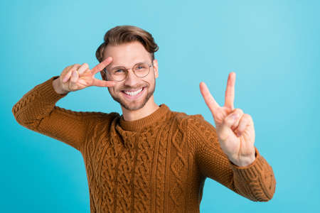 Photo of cheerful charming young man make v-signs wear glasses hello isolated on blue color...
