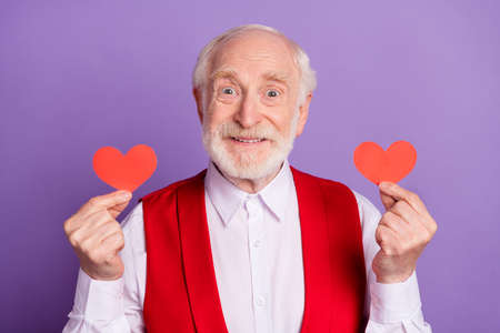 Photo of cheerful old happy positive man hold hands little red paper hearts smile isolated on purple...