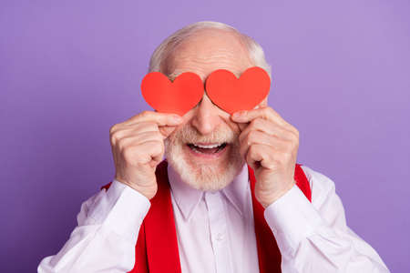 Photo of funky happy positive old man hold hands red paper hearts cover eyes isolated on violet color backgroundの写真素材