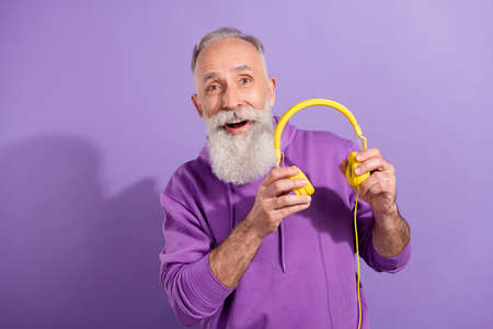 Portrait of attractive cheerful grey-haired man listening music holding earphones stereo isolated...