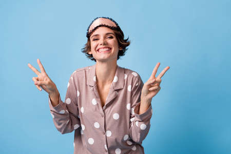 Photo of young attractive happy positive smiling girl wear eye mask and pajamas show v-sign isolated on blue color backgroundの写真素材
