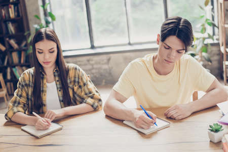 Portrait of two attractive busy focused learners doing home task writing essay at library loft industrial interior indoorsの写真素材