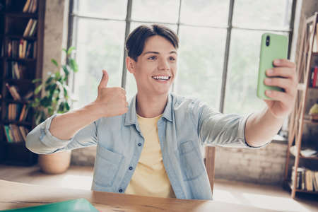 Portrait of attractive cheerful guy pupil making taking selfie showing thumbup having fun at loft industrial interior indoorsの写真素材