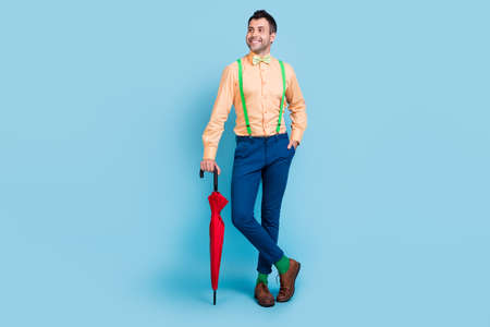 Photo of cheerful guy hold parasol look empty space wear suspenders shirt bow tie isolated blue color backgroundの写真素材