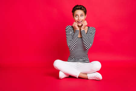 Photo of sweet impressed woman dressed striped outfit legs crossed arms cheeks looking empty space isolated red color backgroundの写真素材