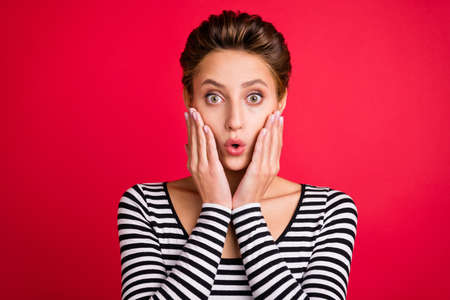 Photo of amazed shocked surprised young woman hold hands face cheekbones isolated on red color backgroundの写真素材