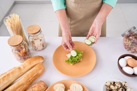 Top angle cropped photo of grandmother prepare homemade meal breakfast put salad lettuce sliced cucumber on bread at homeの写真素材