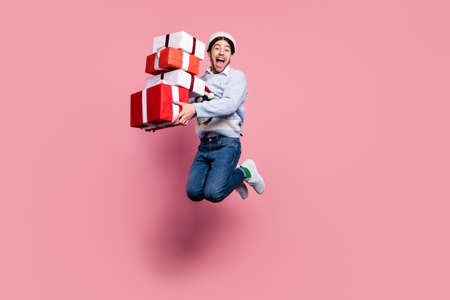 Photo of cute shocked young man dressed blue christmas pullover jumping holding presents pile smiling isolated pink color backgroundの写真素材