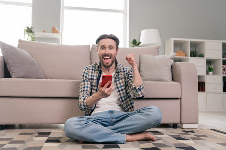 Portrait of attractive lucky cheerful guy sitting on carpet using device having fun staying home at light room indoorsの写真素材