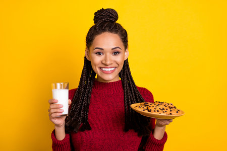 Photo of charming sweet dark skin lady wear red pullover smiling holding cookies milk glass isolated yellow color backgroundの写真素材