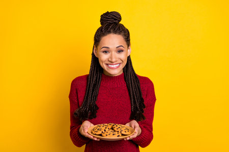 Photo of pretty excited dark skin lady wear red pullover smiling holding cookies plate isolated yellow color backgroundの写真素材