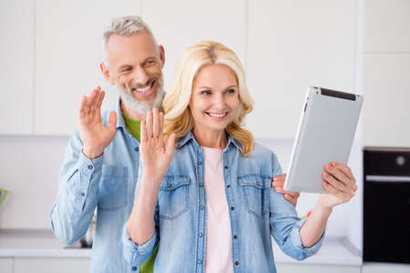 Photo of charming shiny aged couple wear denim shirt smiling waving arms modern device indoors house...