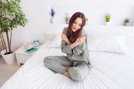 Photo of shiny charming young woman grey nightwear sitting bed hugging herself smiling inside...