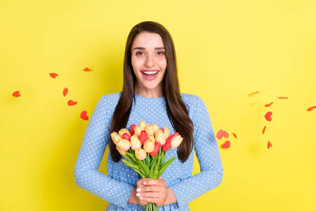 Photo of adorable excited young woman dressed blue clothes holding bouquet smiling isolated yellow color backgroundの写真素材
