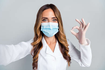 Photo of mature happy woman hold camera make selfie okay sign doctor wear mask isolated on grey color backgroundの写真素材